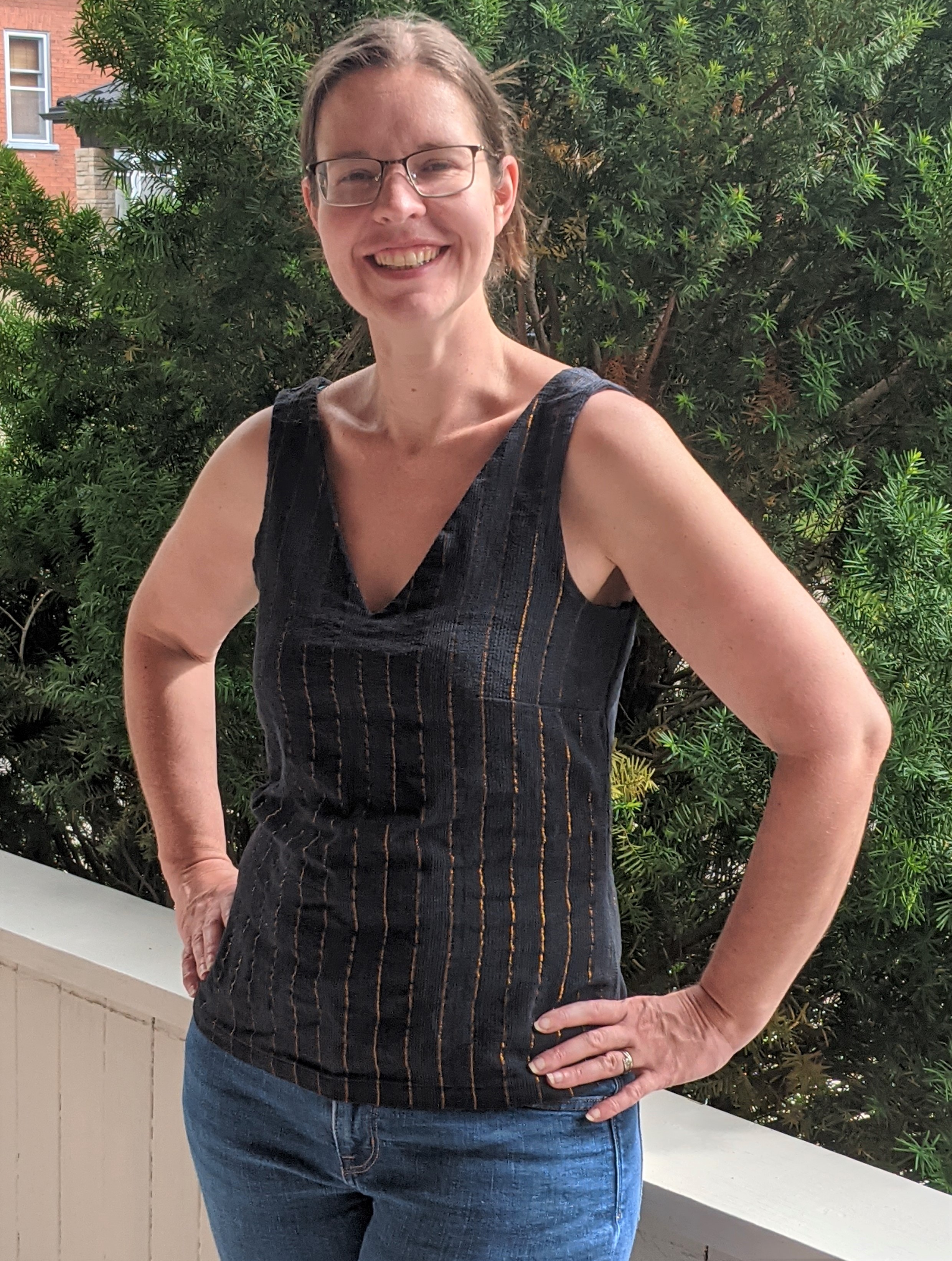 A photo of Jenn standing outside, wearing jeans and a black sleeveless top with bronze sparkly stripes.